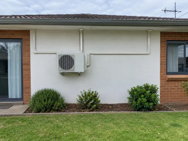 Outdoor air conditioning unit on a house wall with white pipe covers running across the facade in Highbury