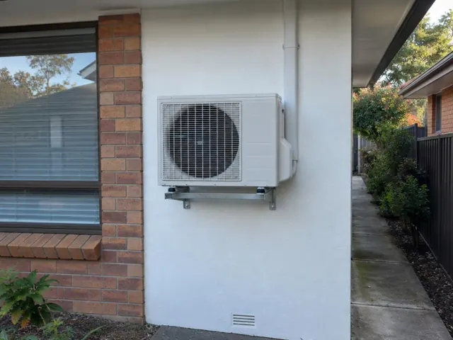 Wall-mounted outdoor air conditioner unit beside a brick home and side path in Highbury